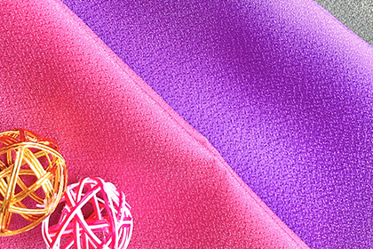 畳んだピンクの風呂敷の上に紫の風呂敷が両方の色見が見えるように少しずれて重なっている。左下には金と赤の水引で作られたボールのようなオブジェがひとつずつ置いてある