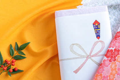 広げた黄色の風呂敷の上に蝶結びの熨斗が巻かれた箱、ピンクの梅柄の風呂敷、反対側には南天の実が乗っている